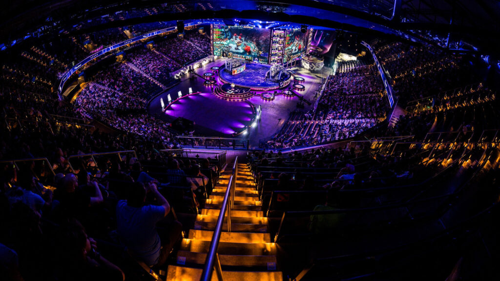 Арена Shanghai Oriental Sports Center заповнена глядачами під час The International 2026. Яскраве освітлення сцени, величезний екран із логотипом турніру, атмосфера напруженого очікування перед стартом матчів. На передньому плані — фанати з прапорами різних країн, серед них помітні символи команд BetBoom Team (BoomBoys) та Team Falcons. Загальна атмосфера — святкове дійство світового масштабу, що підкреслює значення турніру.