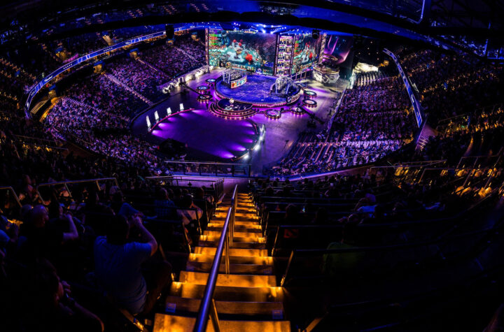 Арена Shanghai Oriental Sports Center заповнена глядачами під час The International 2026. Яскраве освітлення сцени, величезний екран із логотипом турніру, атмосфера напруженого очікування перед стартом матчів. На передньому плані — фанати з прапорами різних країн, серед них помітні символи команд BetBoom Team (BoomBoys) та Team Falcons. Загальна атмосфера — святкове дійство світового масштабу, що підкреслює значення турніру.